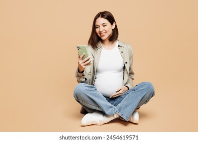 Full body young happy pregnant woman future mom wear grey shirt with belly stomach tummy with baby sitting use mobile cell phone isolated on plain beige background. Maternity family pregnancy concept