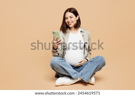 Full body young happy pregnant woman future mom wear grey shirt with belly stomach tummy with baby sitting use mobile cell phone isolated on plain beige background. Maternity family pregnancy concept