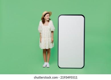 Full Body Young Happy Smiling Woman She 20s In White Dress Hat Look At Big Huge Blank Screen Mobile Cell Phone With Workspace Copy Space Mockup Area Isolated On Plain Pastel Light Green Background.