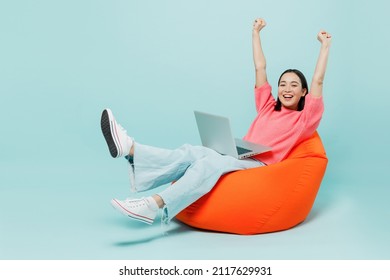 Voller Körper junge glückliche Frau der asiatischen Ethnizität 20er in rosafarbenem Pullover sitzt in Sackstuhl verwenden Arbeit auf Laptop PC mit ausgestreckten Händen beenden Arbeit einzeln auf pastellblauem, hellblauem Hintergrund.