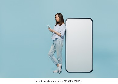 Full body young happy woman she 20s in casual blouse big huge blank screen mobile cell phone with workspace copy space mockup area hold smartphone isolated on pastel plain light blue background studio