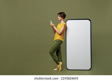 Full Body Young Happy Woman She 20s Wear Yellow T-shirt Near Big Huge Blank Screen Mobile Cell Phone With Workspace Copy Space Mockup Area Use Smartphone Isolated On Plain Olive Green Khaki Background