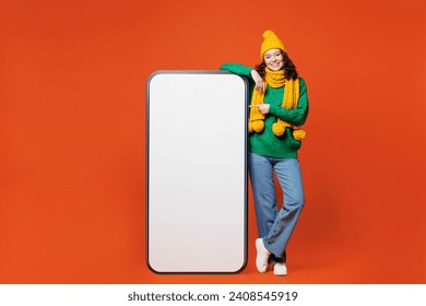 Full body young happy woman she wear green knitted sweater yellow hat scarf big huge point index finger on blank screen mobile cell phone smartphone with area isolated on plain orange red background - Powered by Shutterstock