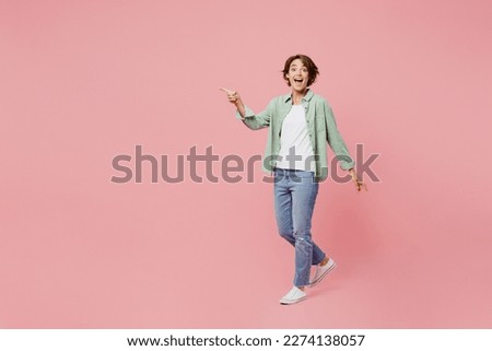 Full body young happy woman 20s she wear green shirt white t-shirt look camera walking going point index finger aside on workspace area isolated on plain pastel light pink background studio portrait