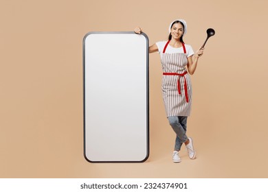 Full body young housewife housekeeper chef baker latin woman wear apron toque hat big huge blank screen mobile cell phone smartphone hold laddle isolated on plain beige background. Cook food concept