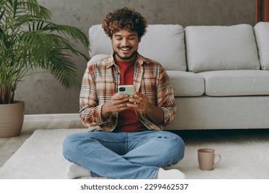 Full body young Indian man wears casual clothes hold in hand use mobile cell phone drink coffee sits near grey sofa couch stay at home hotel flat rest relax spend free spare time in living room indoor - Powered by Shutterstock