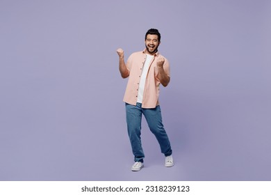 Full body young Indian man he wear pink shirt white t-shirt casual clothes doing winner gesture celebrate clenching fists say yes isolated on plain pastel light purple background. Lifestyle concept