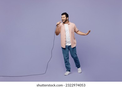 Full body young Indian man he wear pink shirt white t-shirt casual clothes sing song in microphone at karaoke club isolated on plain pastel light purple background studio portrait. Lifestyle concept