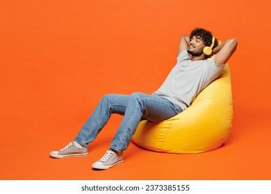 Full body young Indian man wears t-shirt casual clothes sit in bag chair listen to music in headphones hold hands behind neck isolated on orange red color background studio portrait. Lifestyle concept - Powered by Shutterstock