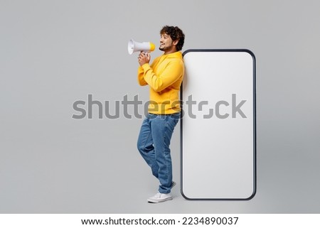 Full body young Indian man wear casual yellow hoody stand near big huge blank screen mobile cell phone with workspace area hold scream in megaphone isolated on plain grey background studio portrait