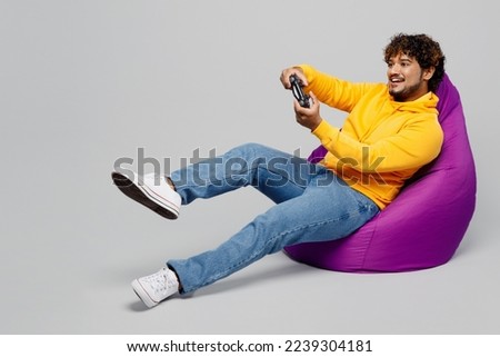 Full body young Indian man he wearing casual yellow hoody sit in bag chair hold in hand play pc game with joystick console isolated on plain grey background studio portrait. People lifestyle portrait