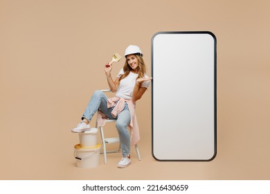 Full body young laborer handyman woman in white t-shirt helmet point blank screen area mobile cell phone hold brush paint isolated on plain beige background Instruments accessories for renovation room - Powered by Shutterstock