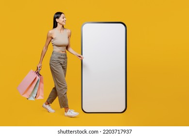 Full Body Young Latin Woman She Wear Tank Shirt Walk Near Big Huge Blank Screen Mobile Cell Phone Workspace Copy Space Holding Package Bags With Shopping Purchases Isolated On Plain Yellow Backround