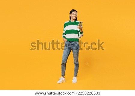 Full body young latin woman wear casual green knitted sweater hold in hand use mobile cell phone chatting online surfing internet isolated on plain yellow background studio People lifestyle concept