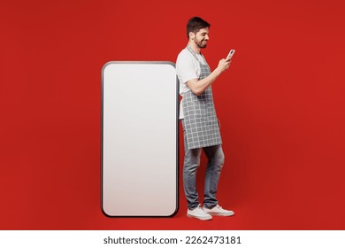 Full body young male housewife housekeeper chef cook baker man wear grey apron big huge blank screen mobile cell phone with area use smartphone isolated on plain red background. Cooking food concept - Powered by Shutterstock