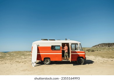 Full body of young male tourist in casual clothes and sunglasses standing in vintage camper van parked on sandy beach before surfing - Powered by Shutterstock