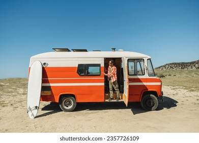 Full body of young male tourist in casual clothes and sunglasses standing in vintage camper van parked on sandy beach before surfing - Powered by Shutterstock