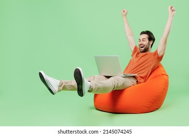 Full body young man 20s wear casual orange t-shirt sit in bag chair hold use work on laptop pc computer stretch hands arms win finish job isolated on plain pastel light green color background studio