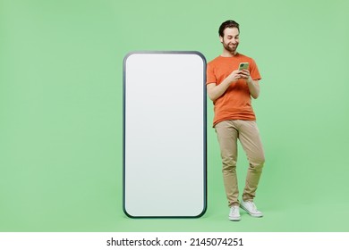 Full Body Young Man 20s Wear Casual Orange T-shirt Stand Uze Mobile Cell Phone With Blank Screen Workspace Area Isolated On Plain Pastel Light Green Color Background Studio. People Lifestyle Concept