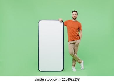 Full body young man 20s wear casual orange t-shirt stand near mobile cell phone with blank screen workspace area isolated on plain pastel light green color background studio. People lifestyle concept