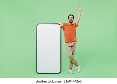 Full Body Young Man 20s Wear Orange T-shirt Stand Near Big Mobile Cell Phone With Blank Screen Workspace Area Greet Meet Isolated On Plain Pastel Light Green Color Background. People Lifestyle Concept