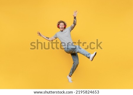 Full body young man 20s he wear grey t-shirt headphones listen to music stand on toes with outstretched hands raise up leg isolated on plain yellow backround studio portrait. People lifestyle concept