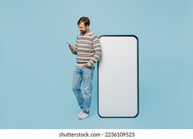 Full body young man 30s wear sweater stand near big huge blank screen mobile cell phone with workspace copy space mockup area use smartphone isolated on plain pastel light blue cyan background studio - Powered by Shutterstock