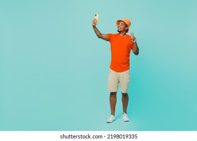 Full body young man of African American ethnicity wear orange t-shirt hat doing selfie shot on mobile cell phone post photo on social network show v-sign isolated on plain pastel light blue background