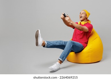 Full body young man of African American ethnicity he wear pink t-shirt yellow hat headphones sit in bag chair hold in hand play pc game with joystick console isolated on plain grey background studio - Powered by Shutterstock