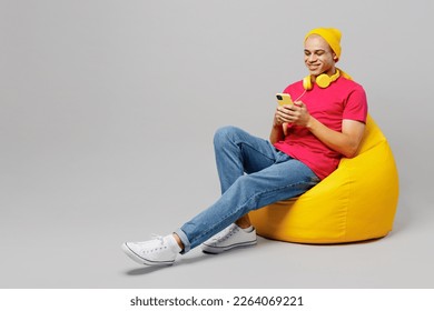 Full body young man of African American ethnicity wear pink t-shirt yellow hat headphones sit in bag chair hold in hand use mobile cell phone chatting online isolated on plain grey background studio - Powered by Shutterstock