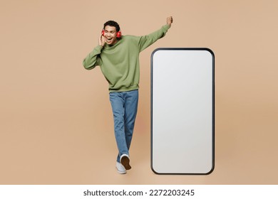 Full body young man of African American ethnicity wear green sweatshirt headohones liste music big huge blank screen mobile cell phone smartphone with area isolated on plain pastel beige background - Powered by Shutterstock