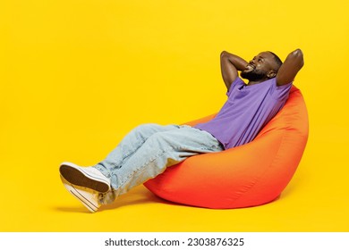 Full body young man of African American ethnicity he wear casual clothes purple t-shirt sit in bag chair hold hands behind neck isolated on plain yellow background studio portrait. Lifestyle concept - Powered by Shutterstock