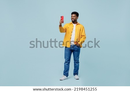 Full body young man of African American ethnicity wear yellow shirt doing selfie shot on mobile cell phone post photo on social network isolated on plain pastel light blue background studio portrait.
