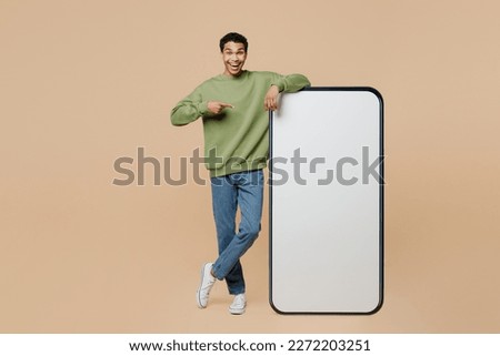 Full body young man of African American ethnicity wears green sweatshirt point index finger on big huge blank screen mobile cell phone smartphone with area isolated on plain pastel beige background