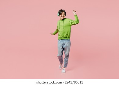 Full body young man of Asian ethnicity wear green hoody headphones listen to music dance laugh have fun raise up hands isolated on plain pastel light pink background studio. People lifestyle concept - Powered by Shutterstock