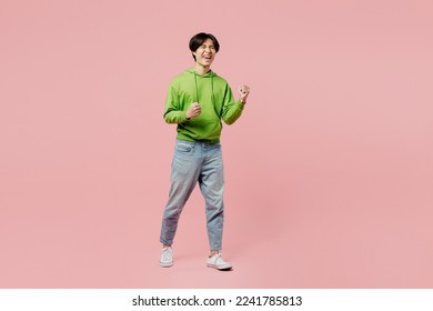 Full body young man of Asian ethnicity wear green hoody doing winner gesture celebrate clenching fists say yes isolated on plain pastel light pink background studio portrait. People lifestyle concept