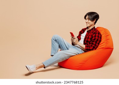 Full body young man of Asian ethnicity he wear red shirt casual clothes sit in bag chair hold in hand use mobile cell phone isolated on plain pastel light beige background studio. Lifestyle concept - Powered by Shutterstock