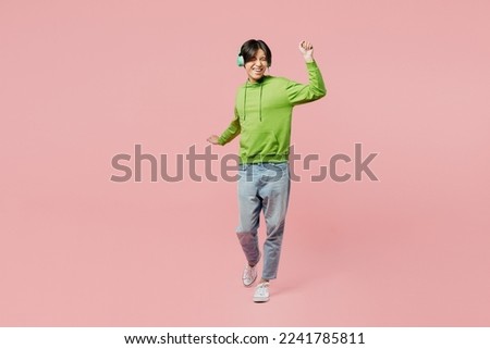 Full body young man of Asian ethnicity wear green hoody headphones listen to music dance laugh have fun raise up hands isolated on plain pastel light pink background studio. People lifestyle concept