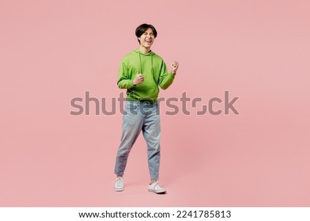 Full body young man of Asian ethnicity wear green hoody doing winner gesture celebrate clenching fists say yes isolated on plain pastel light pink background studio portrait. People lifestyle concept