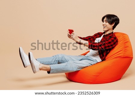 Full body young man of Asian ethnicity wear red shirt casual clothes sit in bag chair play racing app on mobile cell phone hold gadget smartphone for pc video games isolated on plain beige background