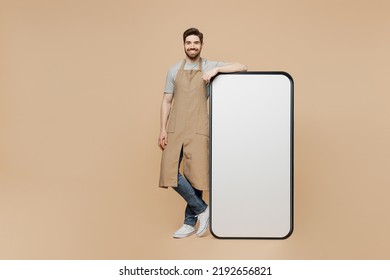 Full Body Young Man Barista Barman Employee Wear Brown Apron Work In Coffee Shop Big Huge Blank Screen Mobile Cell Phone Isolated On Plain Pastel Light Beige Background. Small Business Startup Concept