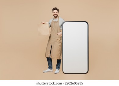 Full Body Young Man Barista Barman Employee Wear Brown Apron Work In Coffee Shop Big Blank Screen Mobile Cell Phone Hold Delivery Bag Isolated On Plain Beige Background. Small Business Startup Concept