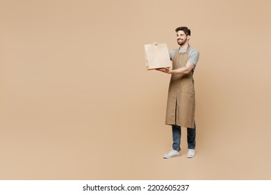 Full Body Young Man Barista Barman Employee Wear Brown Apron Work In Coffee Shop Give Craft Paper Bag For Takeaway Food Isolated On Plain Pastel Light Beige Background. Small Business Startup Concept