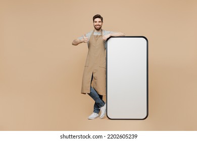 Full Body Young Man Barista Barman Employee In Brown Apron Work In Coffee Shop Huge Blank Screen Mobile Cell Phone Show Like Isolated On Plain Pastel Beige Background Small Business Startup Concept