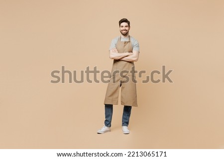 Full body young man barista barman employee wear brown apron work in coffee shop hold hand crossed folded isolated on plain pastel light beige background studio portrait Small business startup concept
