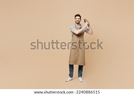Full body young man barista barman employee wear brown apron work in bar pub nightclub using shaker make alcohol cocktail isolated on plain pastel light beige background Small business startup concept
