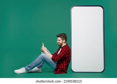 Full Body Young Man He 20s Wear Red Shirt Grey T-shirt Sit Near Big Huge Blank Screen Mobile Cell Phone With Workspace Copy Space Mockup Area Use Smartphone Isolated On Plain Dark Green Background.