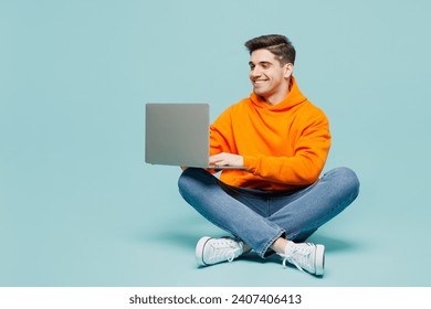 Full body young IT man he wearing orange hoody casual clothes sitting hold use work on laptop pc computer isolated on plain pastel light blue cyan color background studio portrait. Lifestyle concept