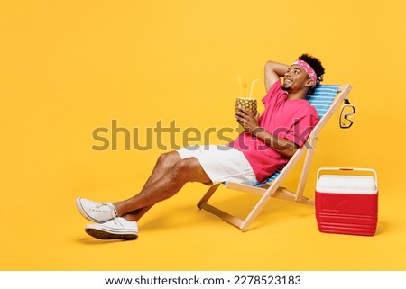 Full body young man he wear pink t-shirt bandana lying on deckchair near hotel pool drink pineapple juice hold hand behind neck isolated on plain yellow background. Summer vacation sea rest concept