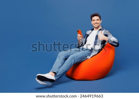 Full body young man he wear shirt white t-shirt casual clothes sit in bag chair hold in hand use point finger on mobile cell phone isolated on plain blue background studio portrait. Lifestyle concept